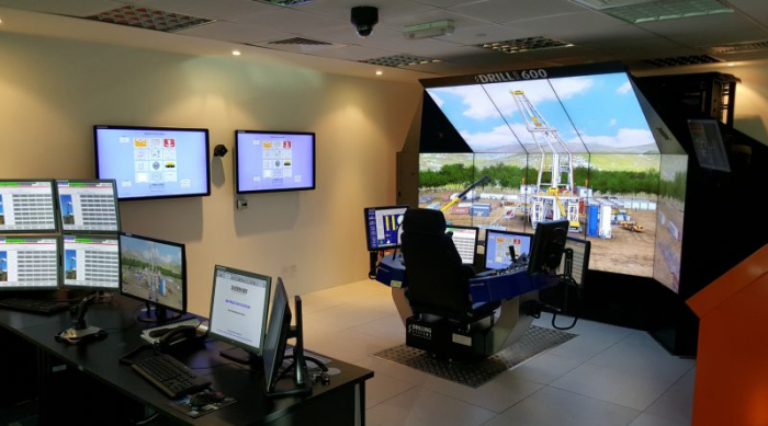 A modern drilling simulator setup with multiple screens showing an outdoor drilling scenario. The control console is equipped with joysticks and monitors displaying various operational data.
