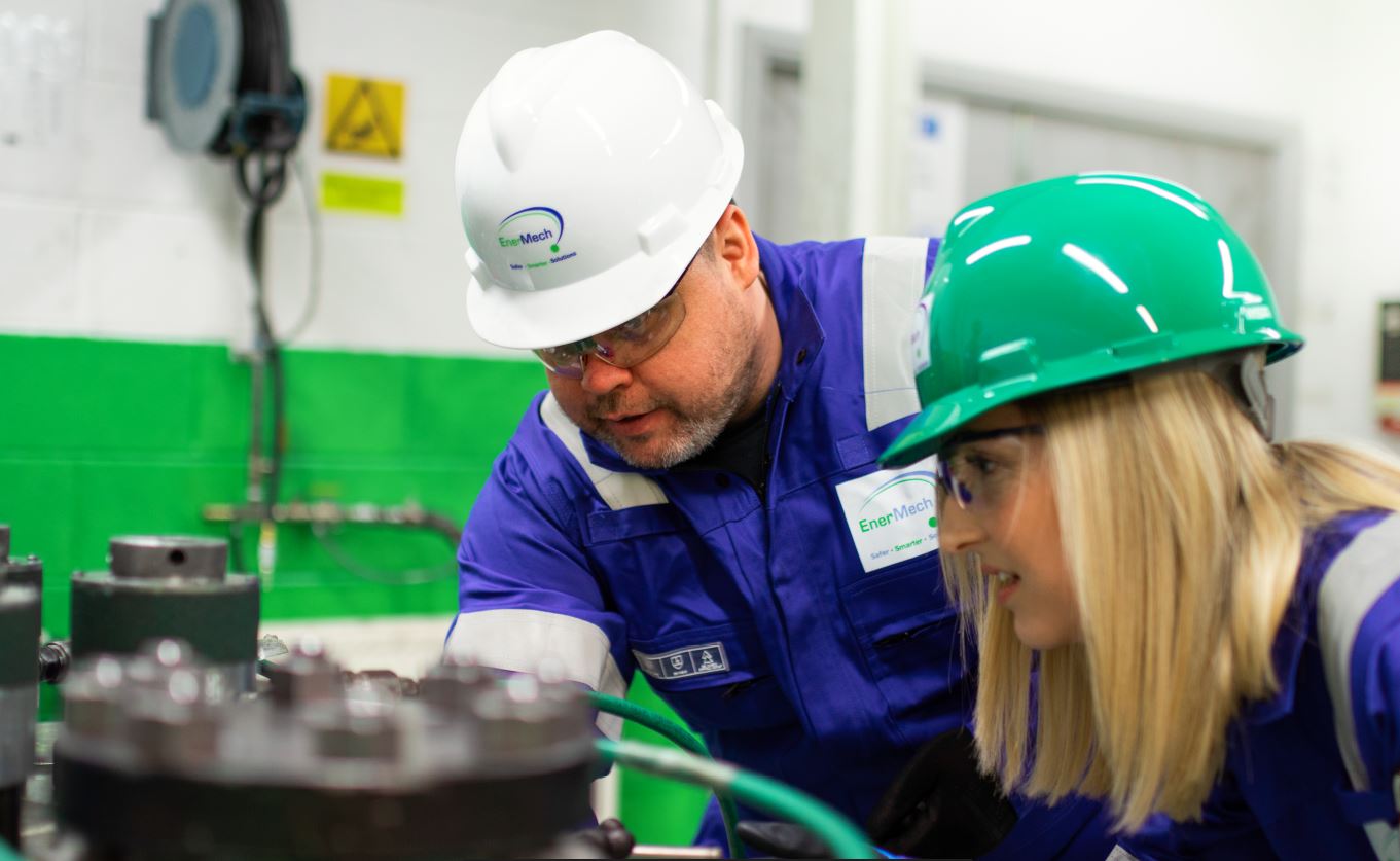 Two workers wearing hard hats and protective clothing inspect machinery in an industrial setting. One worker points out a detail to the other.