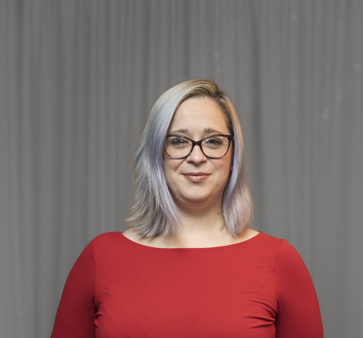 A woman with glasses and shoulder-length light hair is wearing a red top and standing in front of a grey curtain.