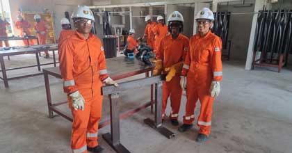 A group of workers in orange coveralls and white helmets stand around a metal structure in an industrial workshop.