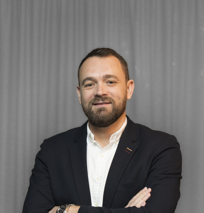 A man with a beard, wearing a navy blazer and white shirt, stands in front of a gray curtain with his arms crossed.