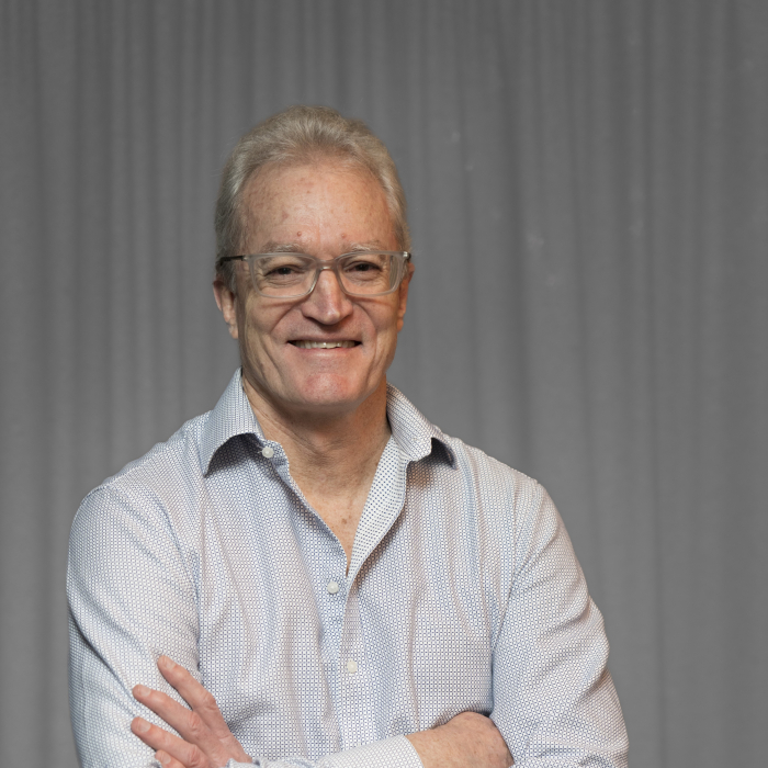 A man with gray hair and glasses is wearing a light blue button-down shirt. He is smiling and standing with his arms folded in front of a gray curtain.
