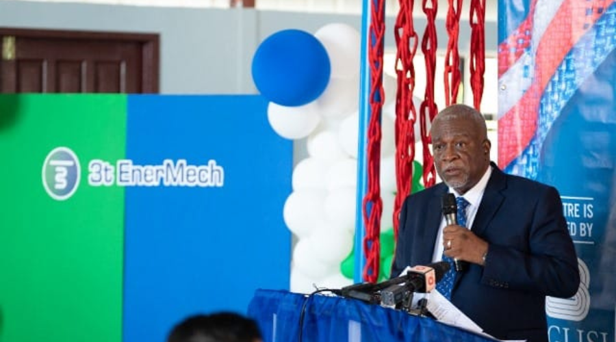 A man in a suit speaks at a podium with a microphone. Behind him are colorful balloons and a 