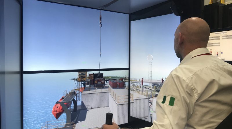 A person operates a crane simulator, controlling a hoist above an offshore platform displayed on large screens.