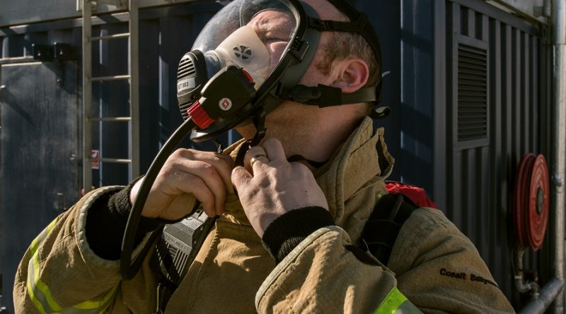 A firefighter adjusts his face mask, wearing protective gear in front of a fire station.
