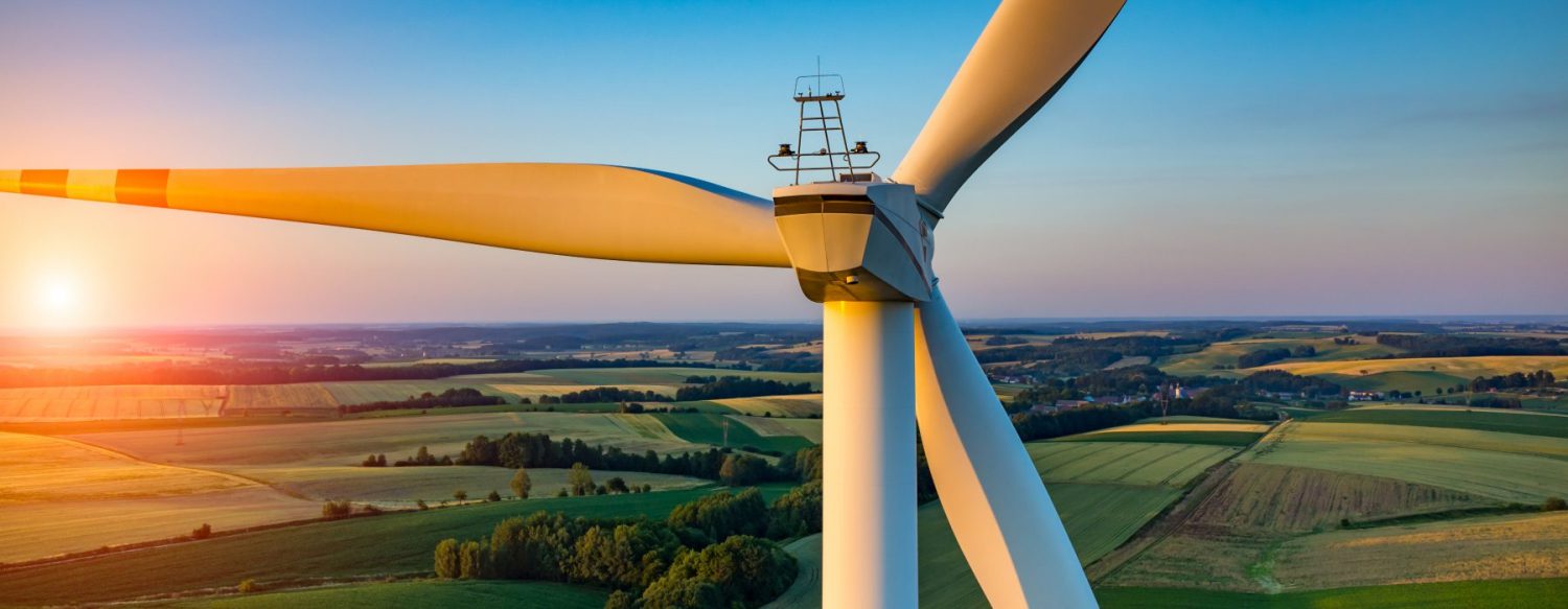 Close-up of a wind turbine with three blades, set against a backdrop of sprawling green fields and a setting sun. The sky is clear with a gradient from blue to orange near the horizon.