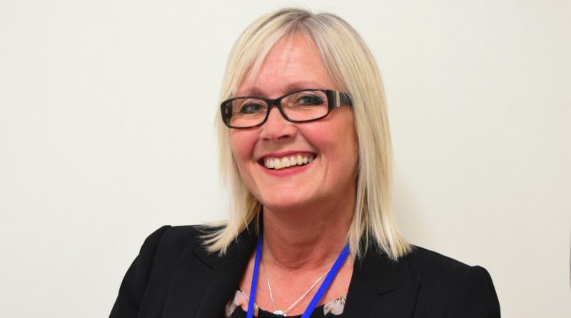 A smiling woman with blonde hair and glasses, wearing a black blazer with a blue lanyard, stands against a plain background.