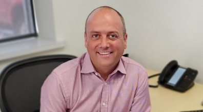 A man in a pink shirt sits at a desk with a phone and window in the background, smiling at the camera.