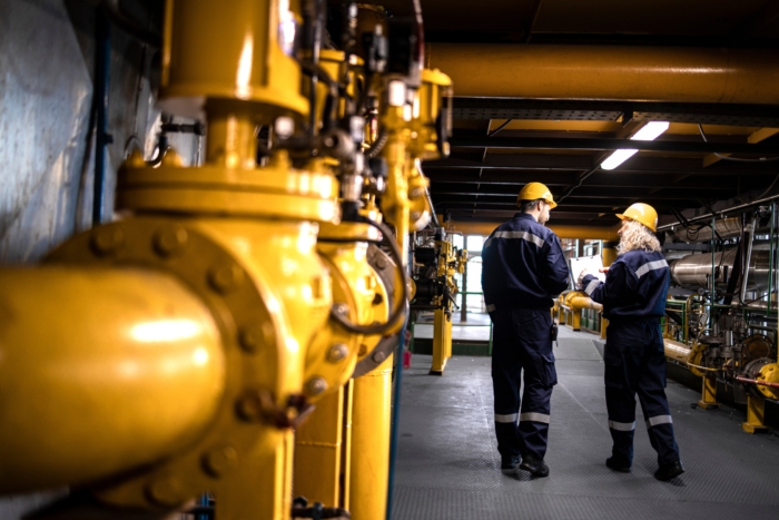 Two engineers in safety gear and helmets analyze documents amid yellow industrial pipes and machinery in a factory setting.