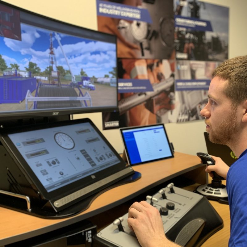 A man operates a control panel while observing a drilling simulation on two screens in a technical training room.