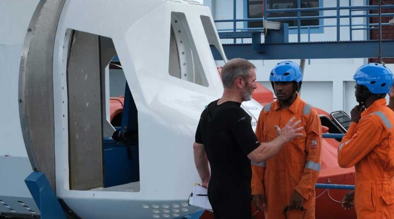 A person in a black shirt instructs two individuals in orange uniforms and blue helmets standing next to a large white apparatus.