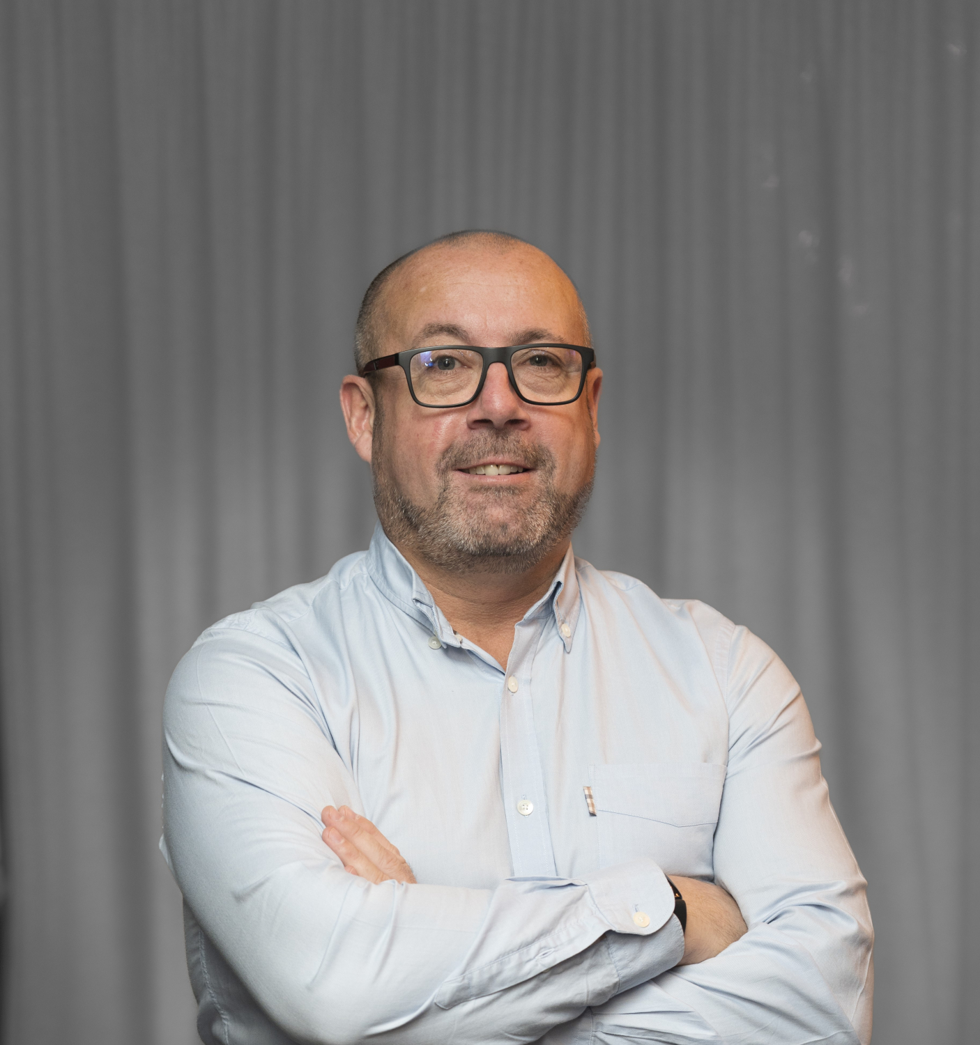 A bald man with glasses and a beard, wearing a light blue button-up shirt, stands with his arms crossed in front of a gray curtain.