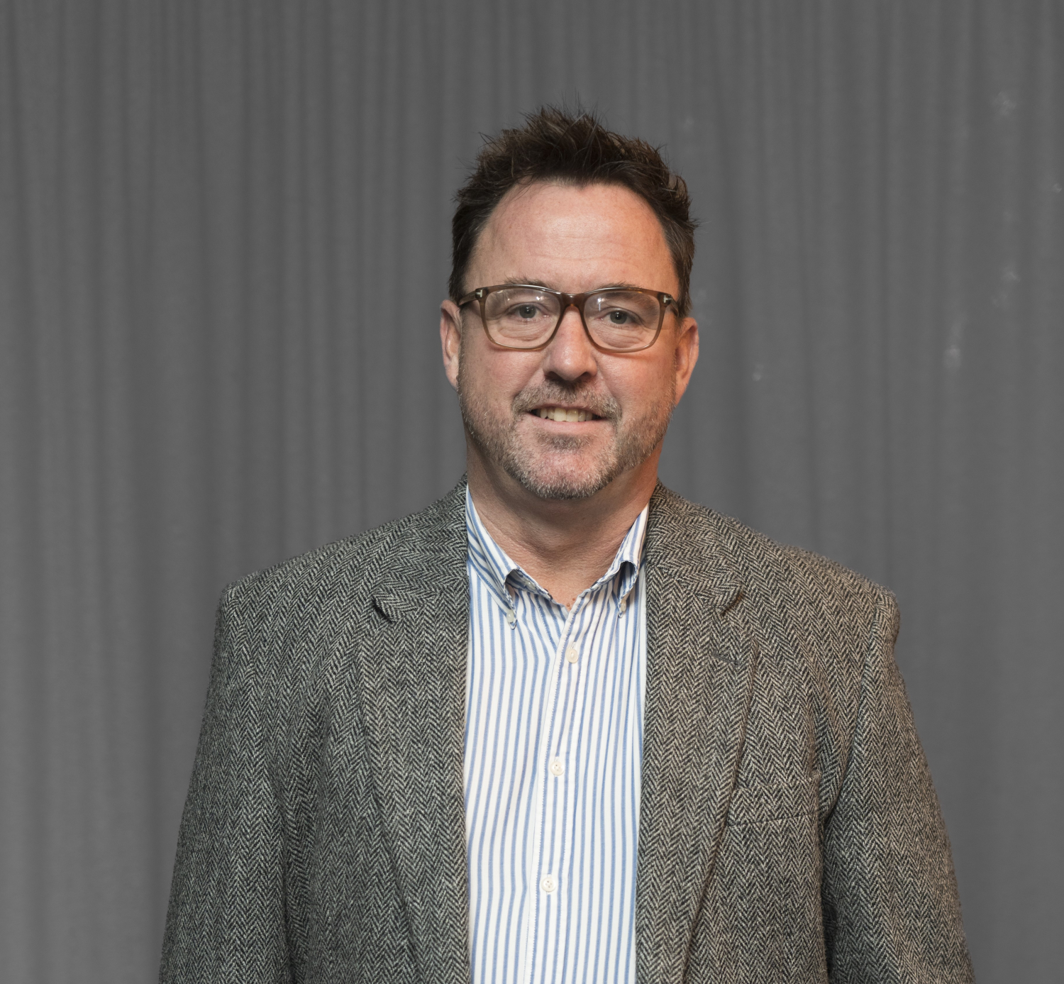 A man with short dark hair, glasses, and a beard is wearing a striped shirt and a tweed blazer, standing in front of a gray curtain.