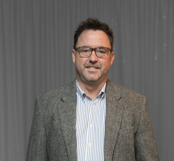 A man with short dark hair, glasses, and a beard is wearing a striped shirt and a tweed blazer, standing in front of a gray curtain.