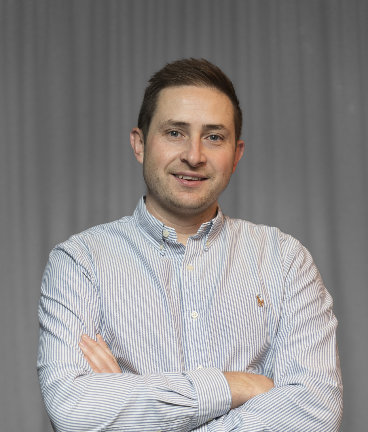 A man with short brown hair wearing a striped button-down shirt stands with arms crossed against a plain backdrop.