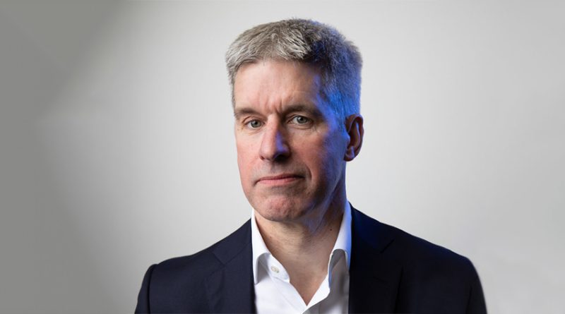 A man with short gray hair in a suit and white shirt poses against a light gray background.