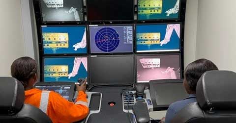 Two seated individuals monitor multiple screens displaying mechanical and technical diagrams in a control room setup. One person operates a joystick controller.