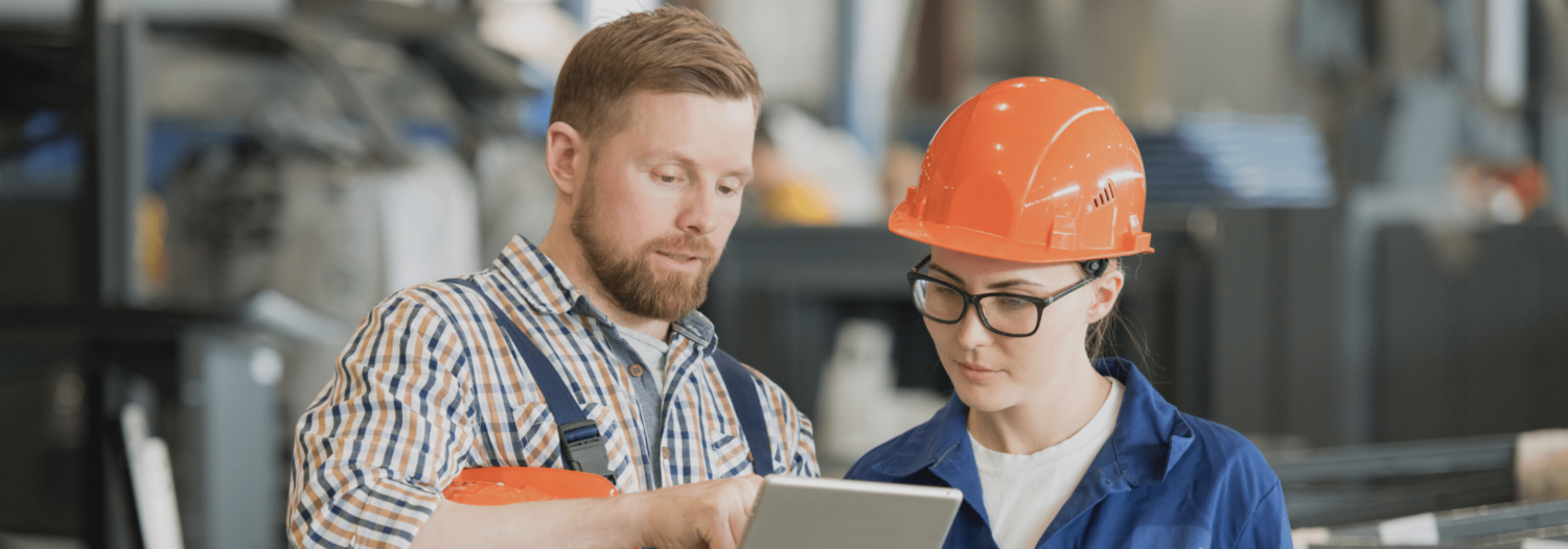 Two workers in a factory setting. One, in plaid shirt, is pointing at a tablet screen, while the other, in a blue uniform and orange hardhat, listens attentively.