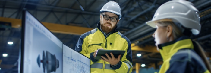 Two workers in high-visibility jackets and hard hats are reviewing information on a tablet and a large screen in an industrial setting.