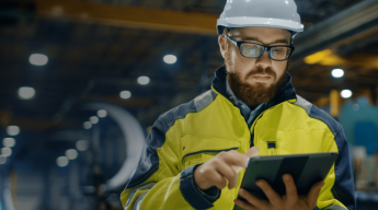 Man in high-visibility jacket and hard hat uses a tablet in an industrial setting.