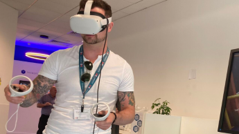 A person wearing a white VR headset and holding controllers stands in a room with white walls and a television screen. They wear a white t-shirt and a lanyard, and have tattoos on their left arm.