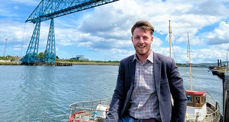 A man in a blazer stands on a boat with a river and a large blue industrial structure in the background under a partly cloudy sky.