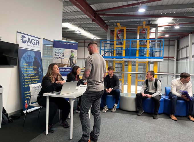 People engaging in conversation at a table in an indoor industrial setting, with company banners displayed in the background.