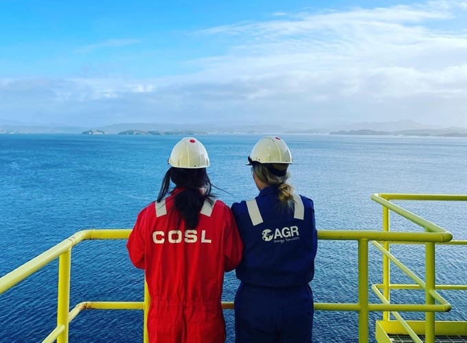 Two workers in helmets and coveralls, labeled COSL and AGR, stand on an offshore platform railing, overlooking a vast expanse of blue sea and distant land.