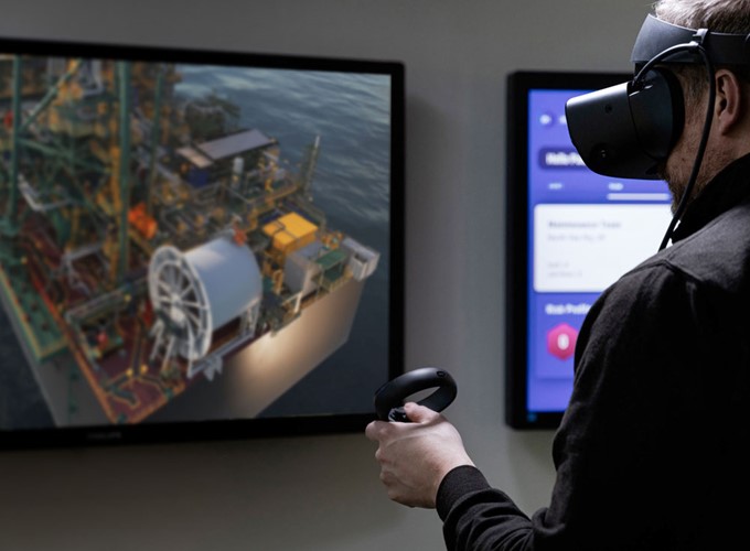 A person wearing a VR headset and holding a controller is viewing a detailed 3D model of an industrial facility displayed on a screen.