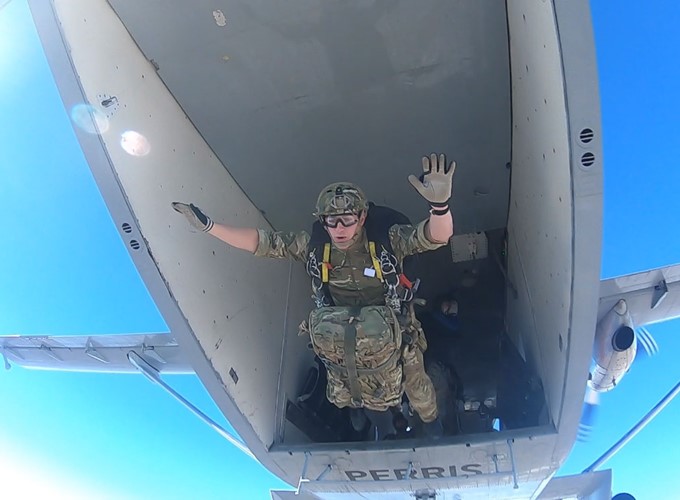 A soldier in full gear, including a parachute, exits from the rear of an aircraft during a skydive, seen waving with one hand, with a clear blue sky in the background.