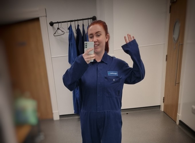 A person with red hair is taking a mirror selfie in a room, wearing a blue jumpsuit and holding a smartphone. There are hanging blue jumpsuits in the background.