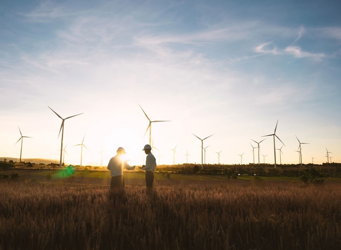 Two people standing in a field at sunset with several wind turbines in the background.
