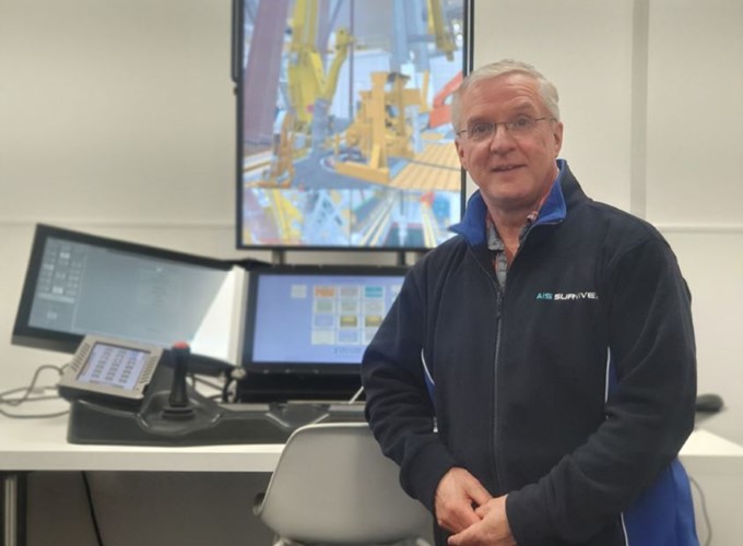 A man wearing glasses and a navy jacket stands in front of a control station with multiple screens displaying industrial equipment.