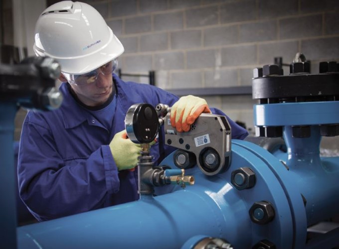 A worker in a blue jumpsuit and white helmet uses equipment to perform maintenance on blue industrial piping in a facility with a concrete block wall.