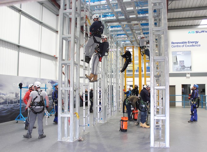 People in safety gear and helmets practice climbing on a metal structure in an indoor training center labeled as the Renewable Energy Centre of Excellence.