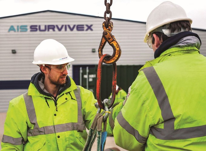 Two workers in high-visibility jackets and hard hats inspect a suspended hook. A building with the sign 