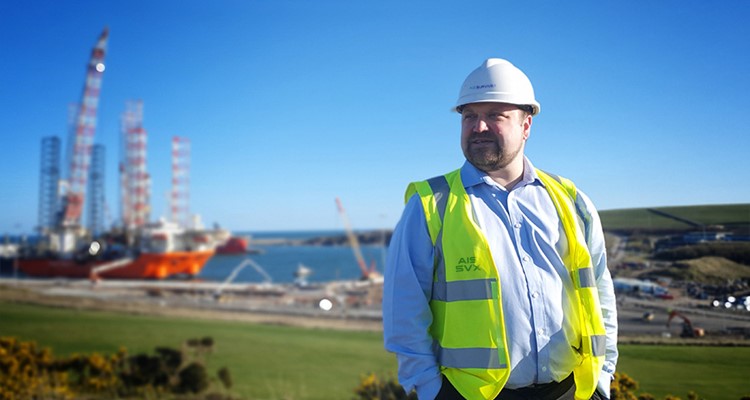 A man wearing a white hard hat and a high-visibility vest stands outdoors with a coastal construction site and cranes in the background.