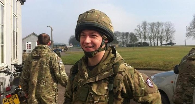 A person in military uniform smiles outdoors near a building and a parked motorcycle, with other uniformed individuals and trees in the background.