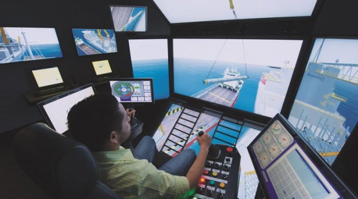 A person operates a complex maritime simulator with multiple screens, showing a ship being loaded. Various control panels and monitors are visible in the simulator room.