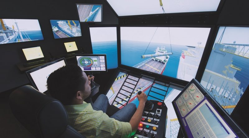 A person operates a complex maritime simulator with multiple screens, showing a ship being loaded. Various control panels and monitors are visible in the simulator room.
