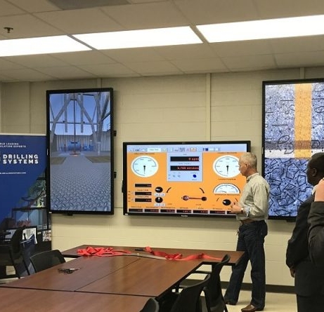 Three men observe multiple digital displays featuring industrial drilling systems in a control room setting. A conference table with red ribbons is in the foreground.