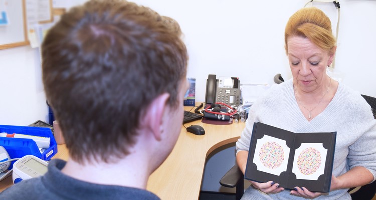 A healthcare professional holds and discusses an Ishihara color blindness test booklet with a patient in an office setting.