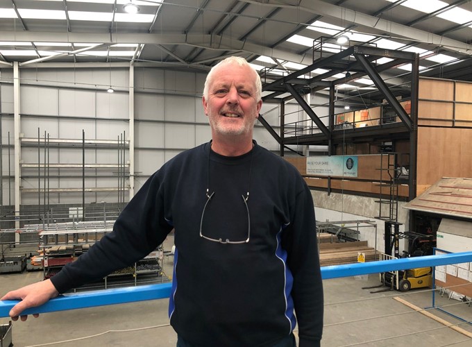A man with a grey beard and a dark sweater stands inside an industrial warehouse, smiling at the camera.