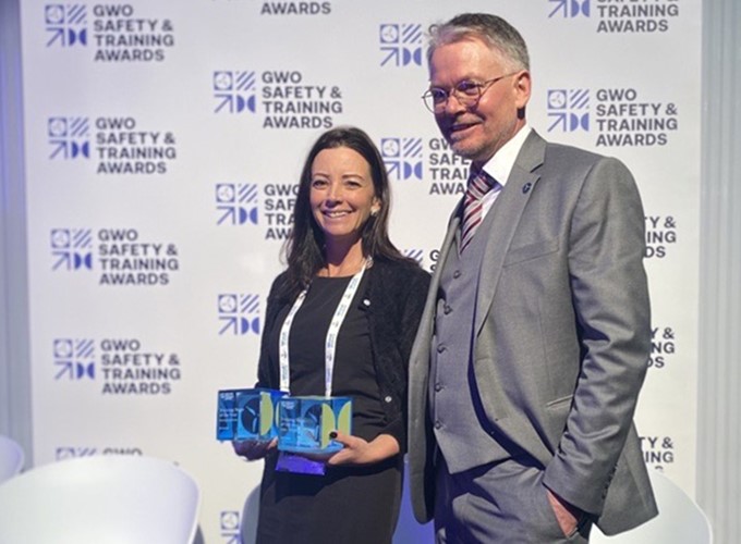 Two individuals stand together at the GWO Safety & Training Awards event. The woman holds two awards, and both are smiling. The backdrop displays the event's logo repeatedly.