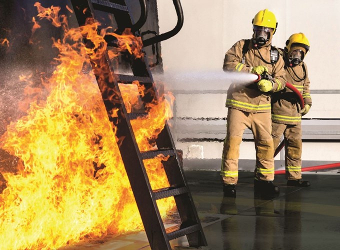 Two firefighters in full gear, including helmets and breathing apparatus, extinguishing a large blaze with a hose on an outdoor metal structure.