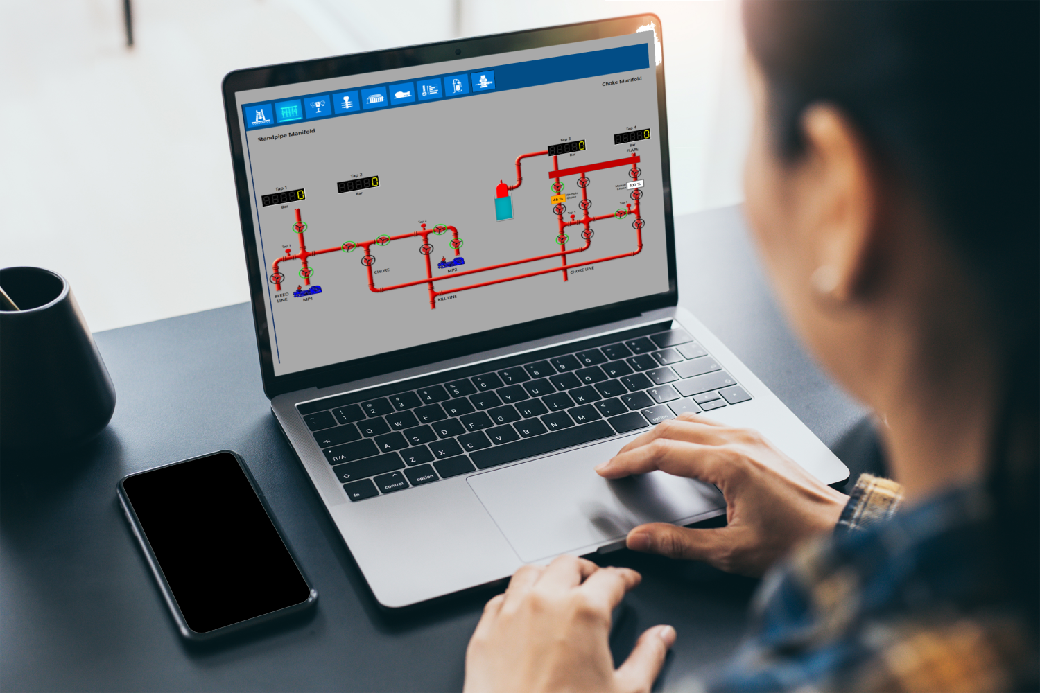 A person operates a laptop displaying a schematic diagram of a technical system with piping and instrumentation. A smartphone and a cup are on the desk.