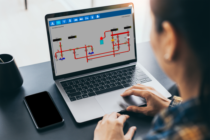 A person operates a laptop displaying a schematic diagram of a technical system with piping and instrumentation. A smartphone and a cup are on the desk.