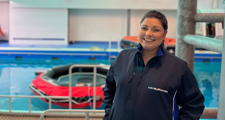 A person in a navy blue jacket stands smiling in front of a pool equipped with lifeboats and safety equipment.