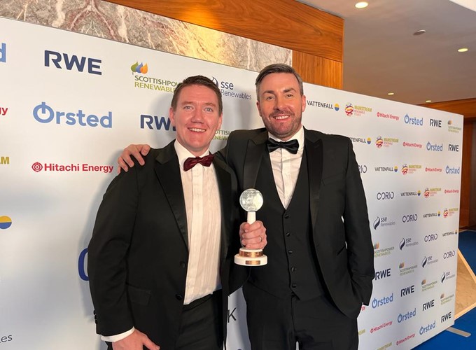 Two men in tuxedos stand on a red carpet. One holds an award trophy. The backdrop features logos of various sponsors.
