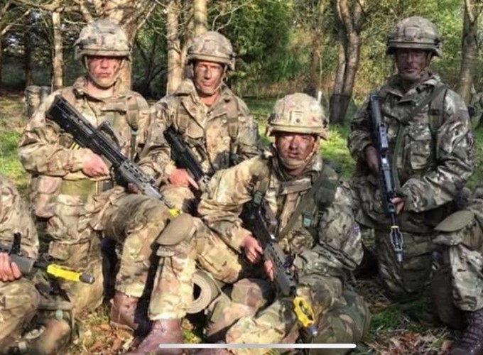 A group of soldiers in camouflage uniforms and helmets pose for a photo in a forested area, holding rifles with yellow attachments.